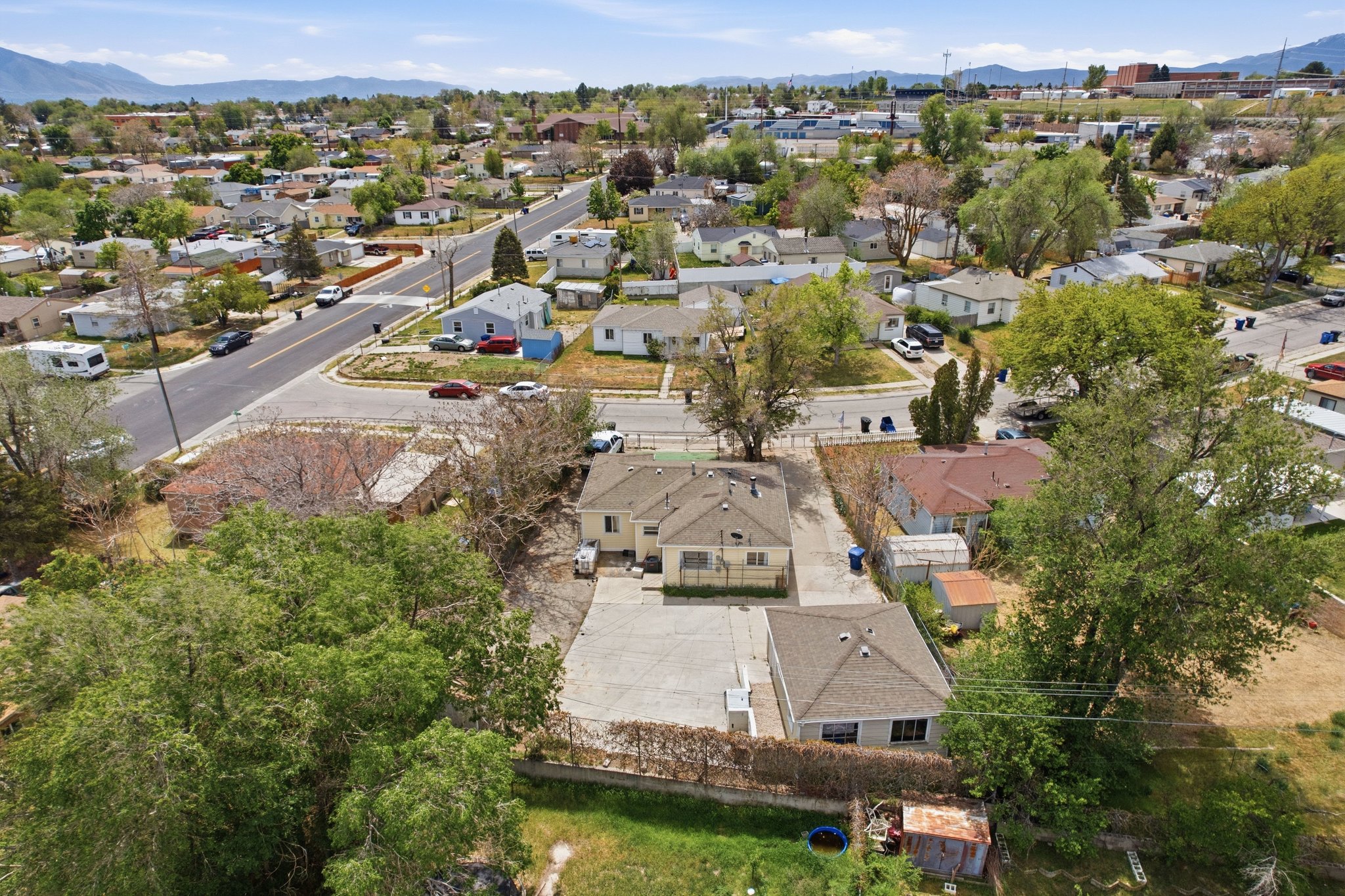 Aerial view showcasing a residential property with a detached accessory structure, expansive paved area, mature trees, and a distant mountain range backdrop