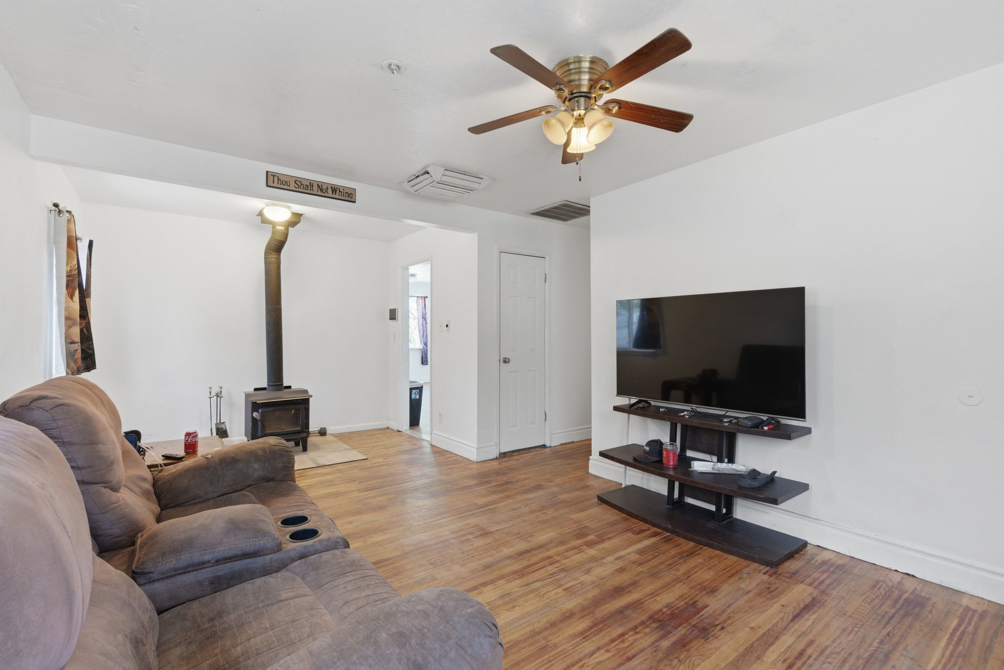 Living area featuring wood-finish flooring, a ceiling fan with light fixture, white painted walls, a wood-burning stove with metal flue, and white trim