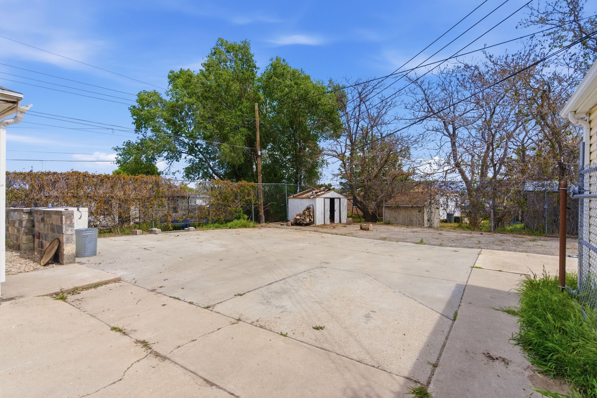 Paved concrete yard area featuring a detached storage shed, a chain-link fence, and mature perimeter trees