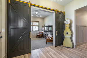 Dark-paneled barn doors with gold-finish hardware open to a room featuring gray carpeting, a white ceiling fan, and window blinds