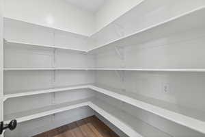 Spacious walk-in pantry featuring white shelving, a wood-finish floor, and an electrical outlet