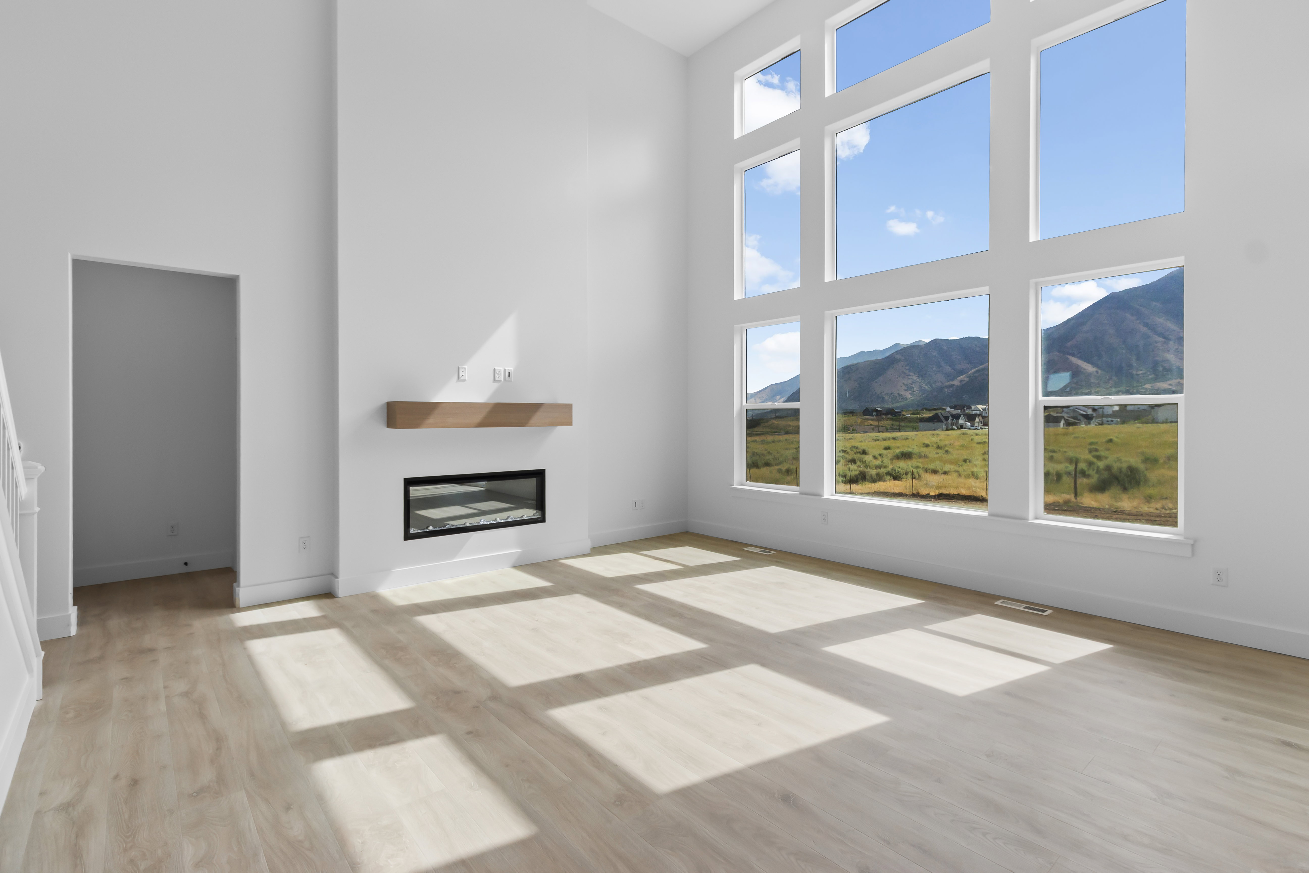 Two-story living area featuring tall windows with mountain scenery, light wood-finish flooring, a modern electric fireplace with a wood mantel, and white walls
