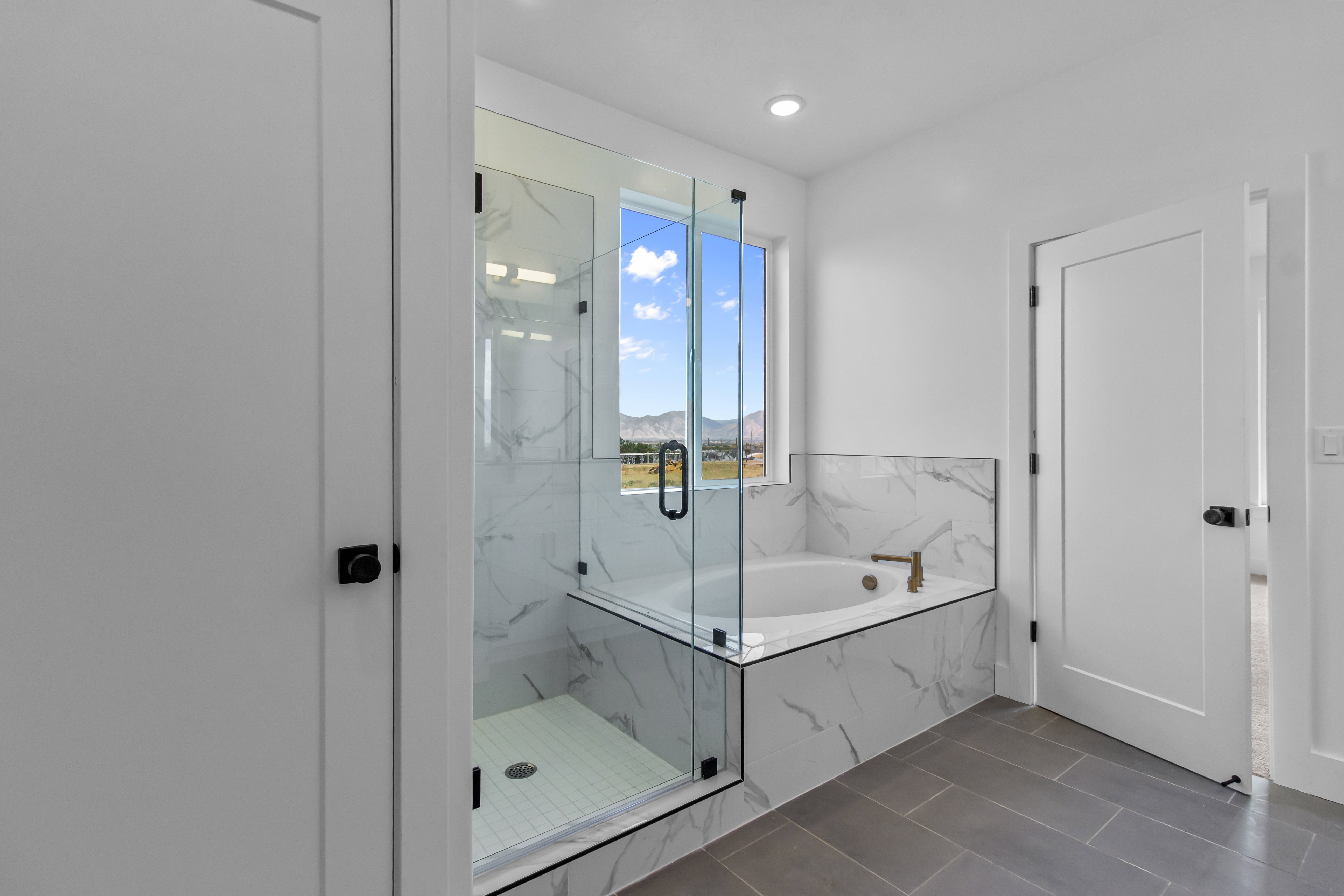 Modern bathroom featuring a walk-in glass shower enclosure, a built-in soaking tub, a large window offering expansive mountain scenery, large format tile flooring, and marble-look wall tile accents