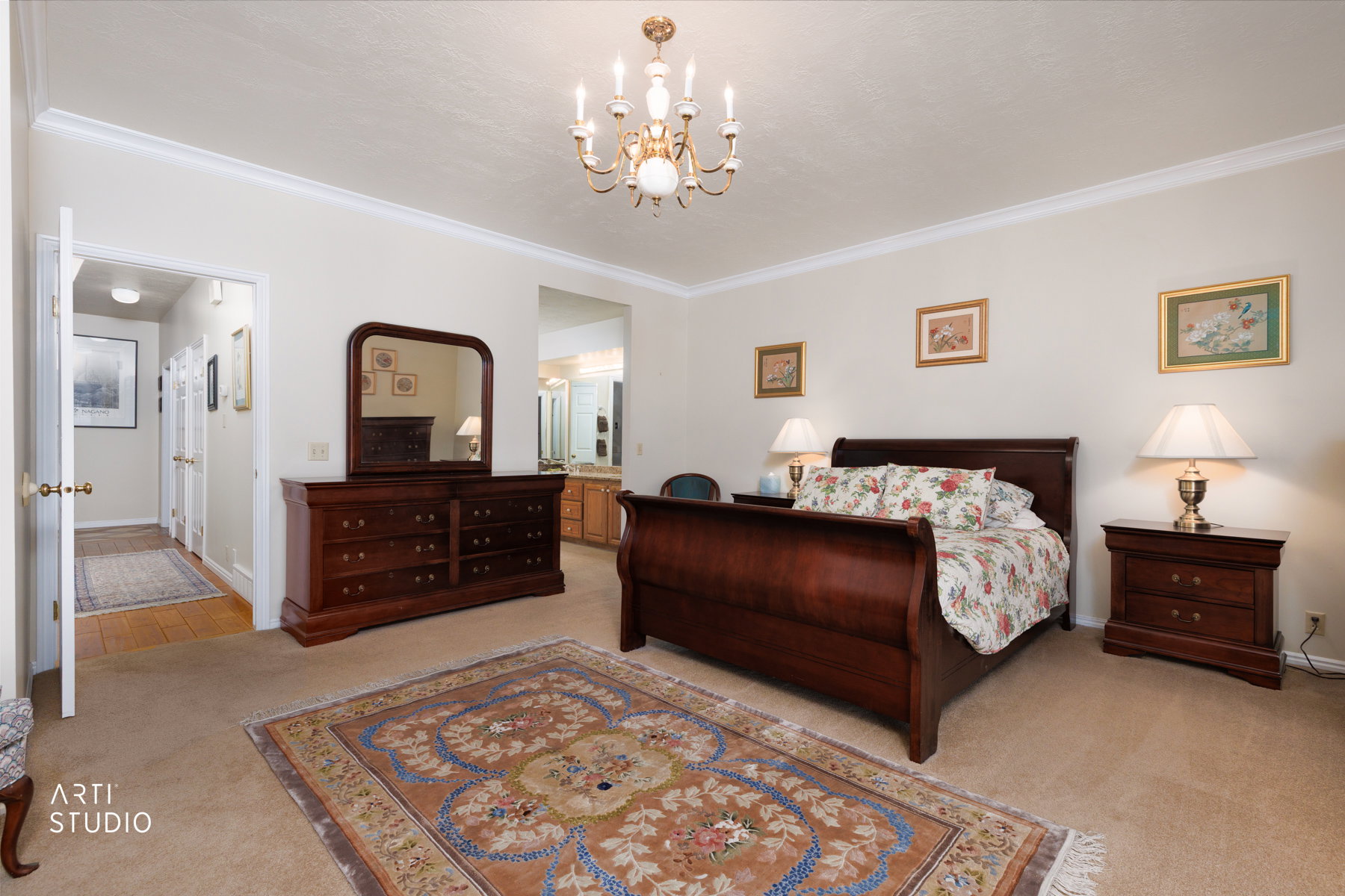 Carpeted  master bedroom featuring a ceiling-mounted chandelier, crown molding, and an open doorway revealing a bathroom with dual vanities