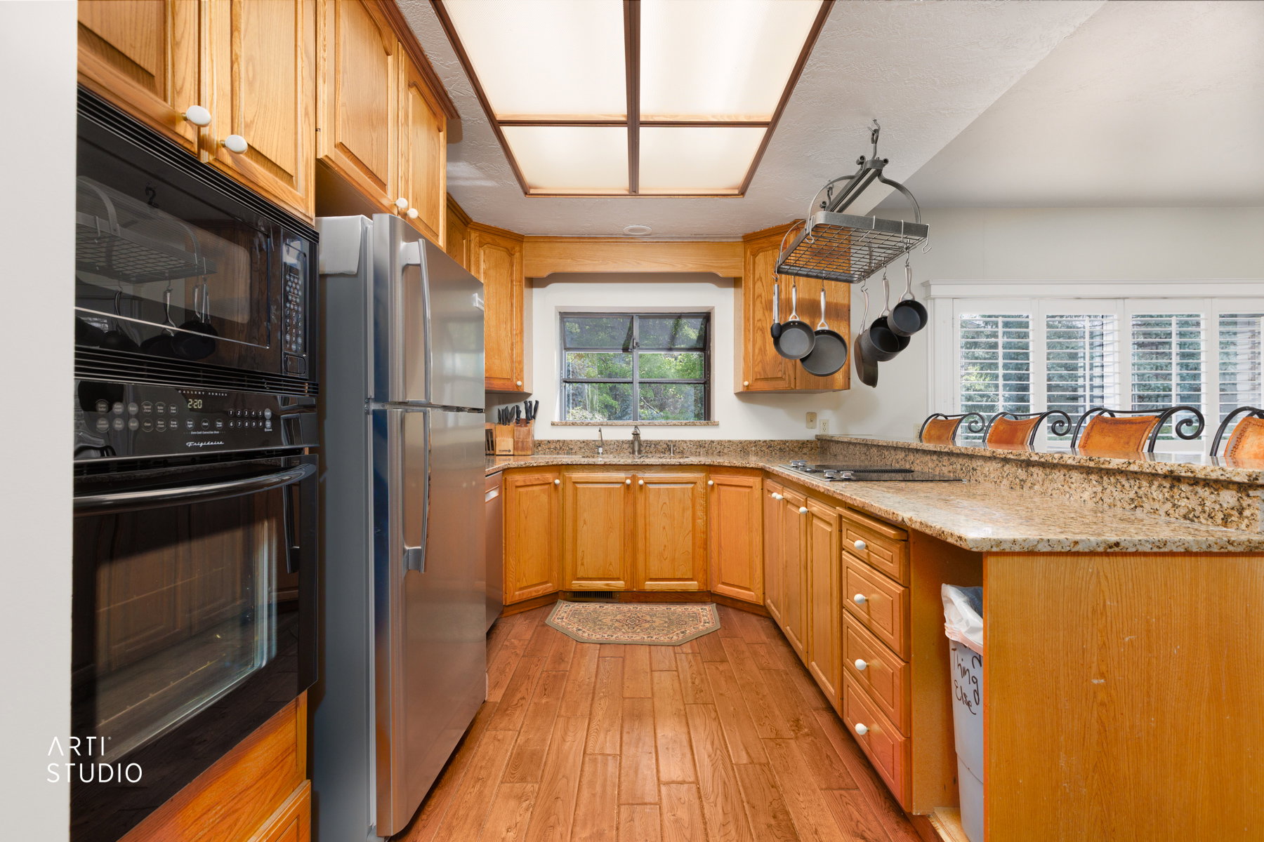 Kitchen featuring wood-finish flooring, light wood cabinetry, stainless steel appliances, granite countertops, and a full window with plantation shutters
