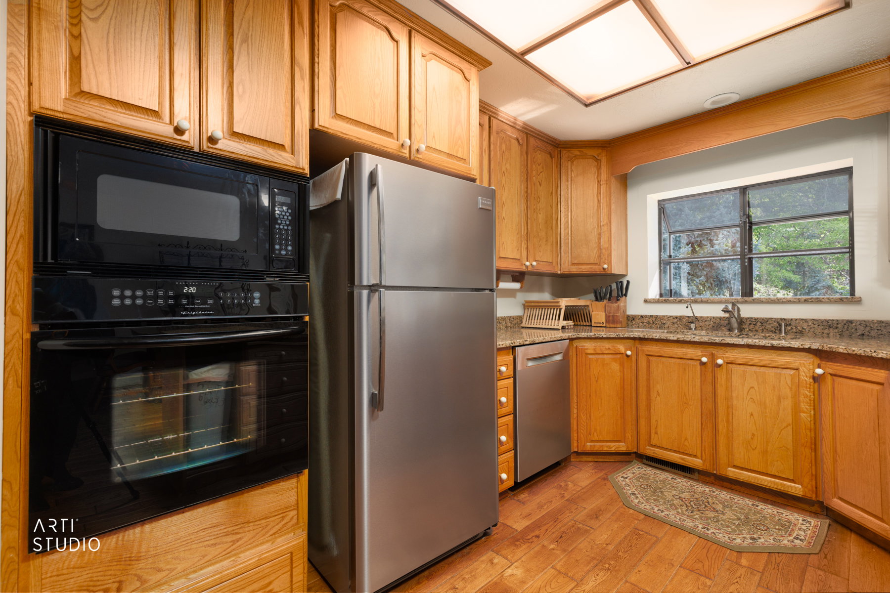 Kitchen featuring wood-finish flooring, light wood cabinetry, stainless steel refrigerator and dishwasher, black wall oven and microwave, and a window above the sink