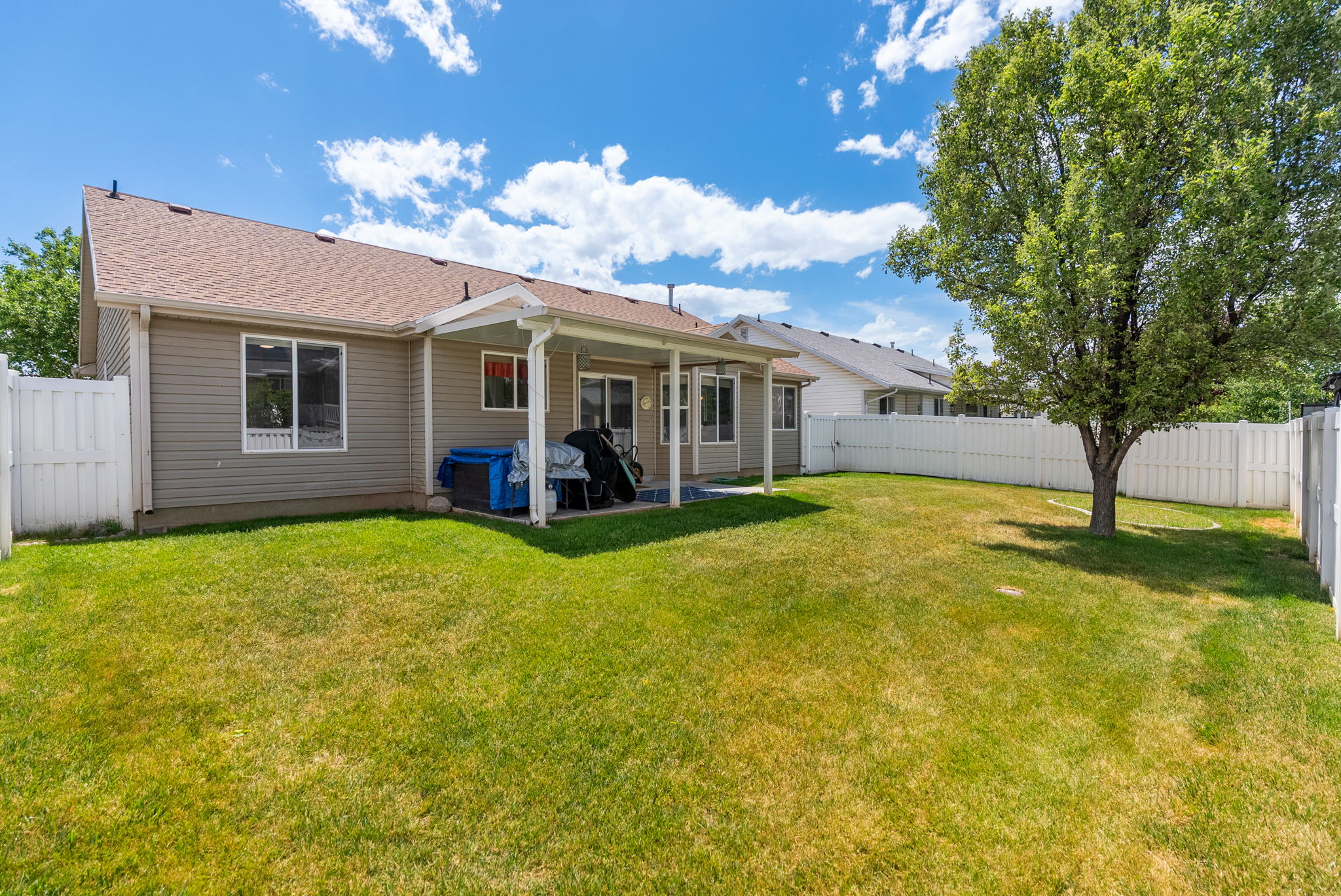 Expansive backyard featuring a manicured lawn, a mature shade tree, a white privacy fence, and a covered patio area
