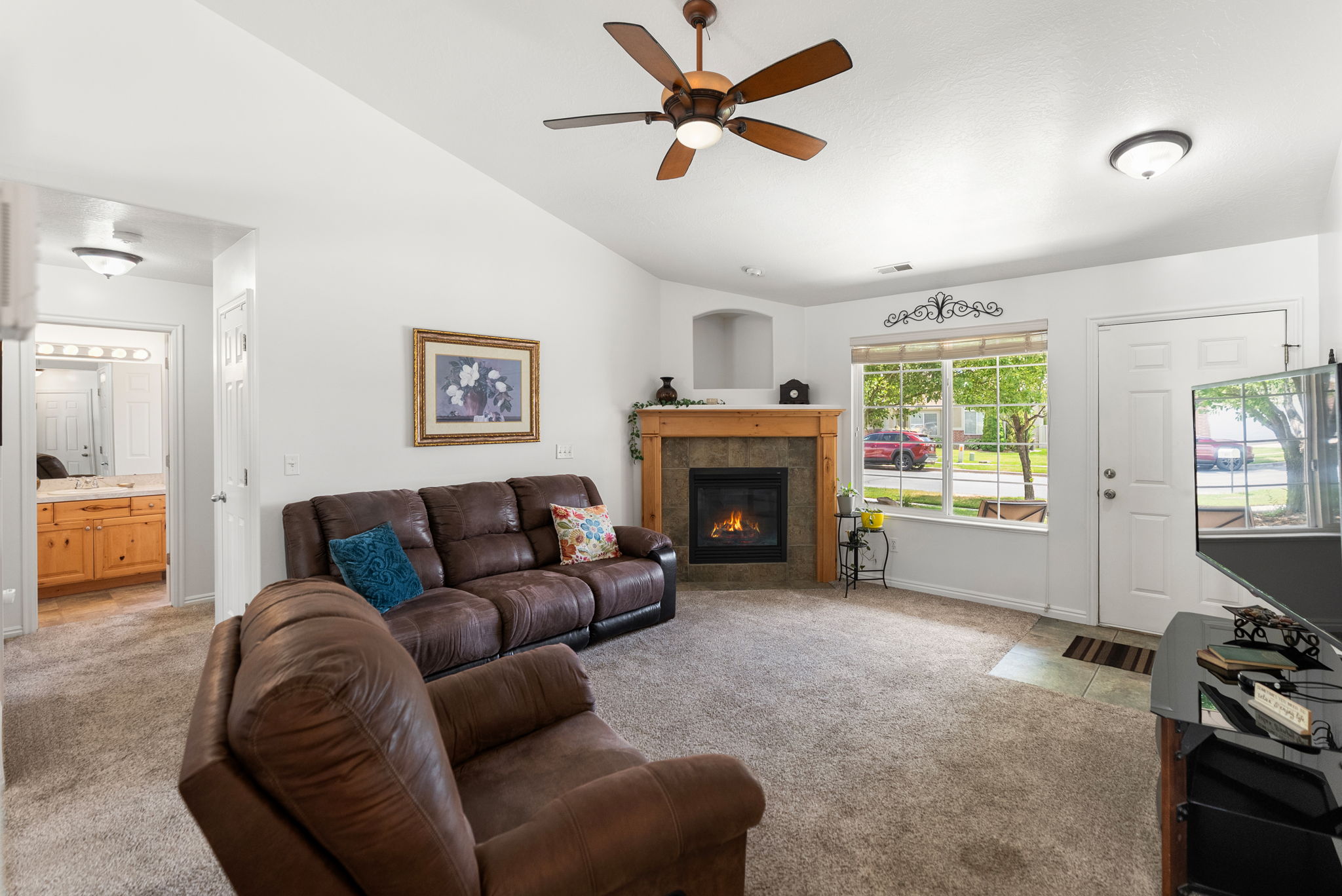 Living area featuring vaulted ceilings, a ceiling fan with integrated lighting, a corner fireplace with a stone surround and wood mantel, large windows with exterior views, and neutral carpet flooring