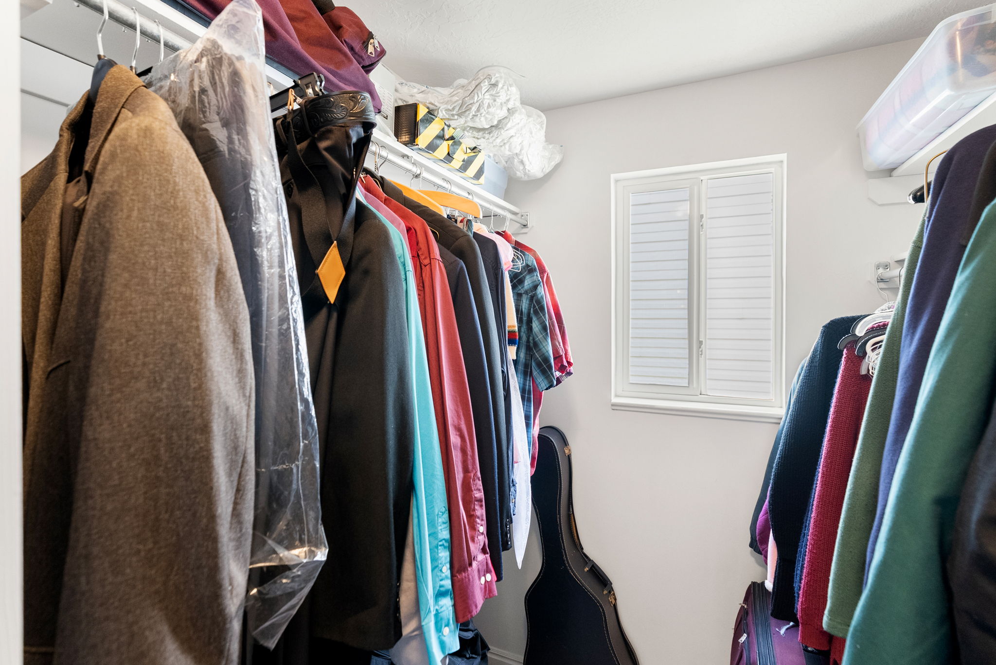 Wardrobe space featuring a white frame window with louvered shutters, light-colored walls, and built-in closet shelving