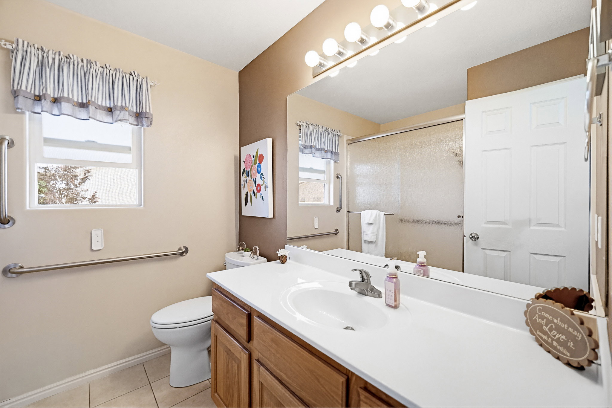 Bathroom featuring a wood-finish vanity with a white countertop and integrated sink, a large mirror with a vanity light bar, a window with natural light, and a shower with a sliding glass enclosure