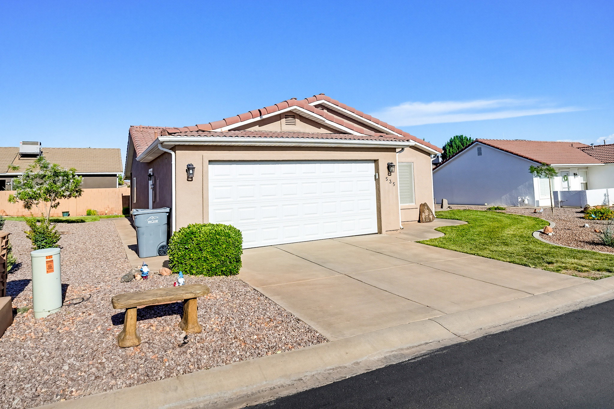 Stucco exterior with a terracotta tile roof, attached garage, concrete driveway, and rock landscaping with mature shrubs