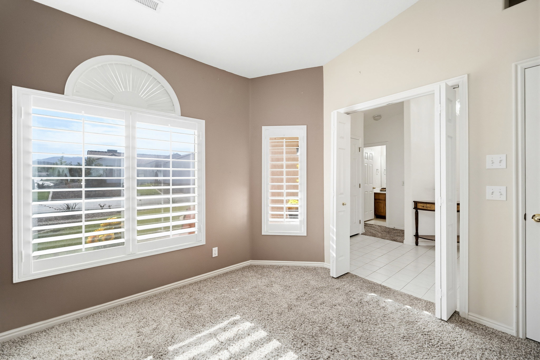 Carpeted room featuring a large arched window with plantation shutters, additional window with plantation shutters, neutral painted walls, and an open doorway