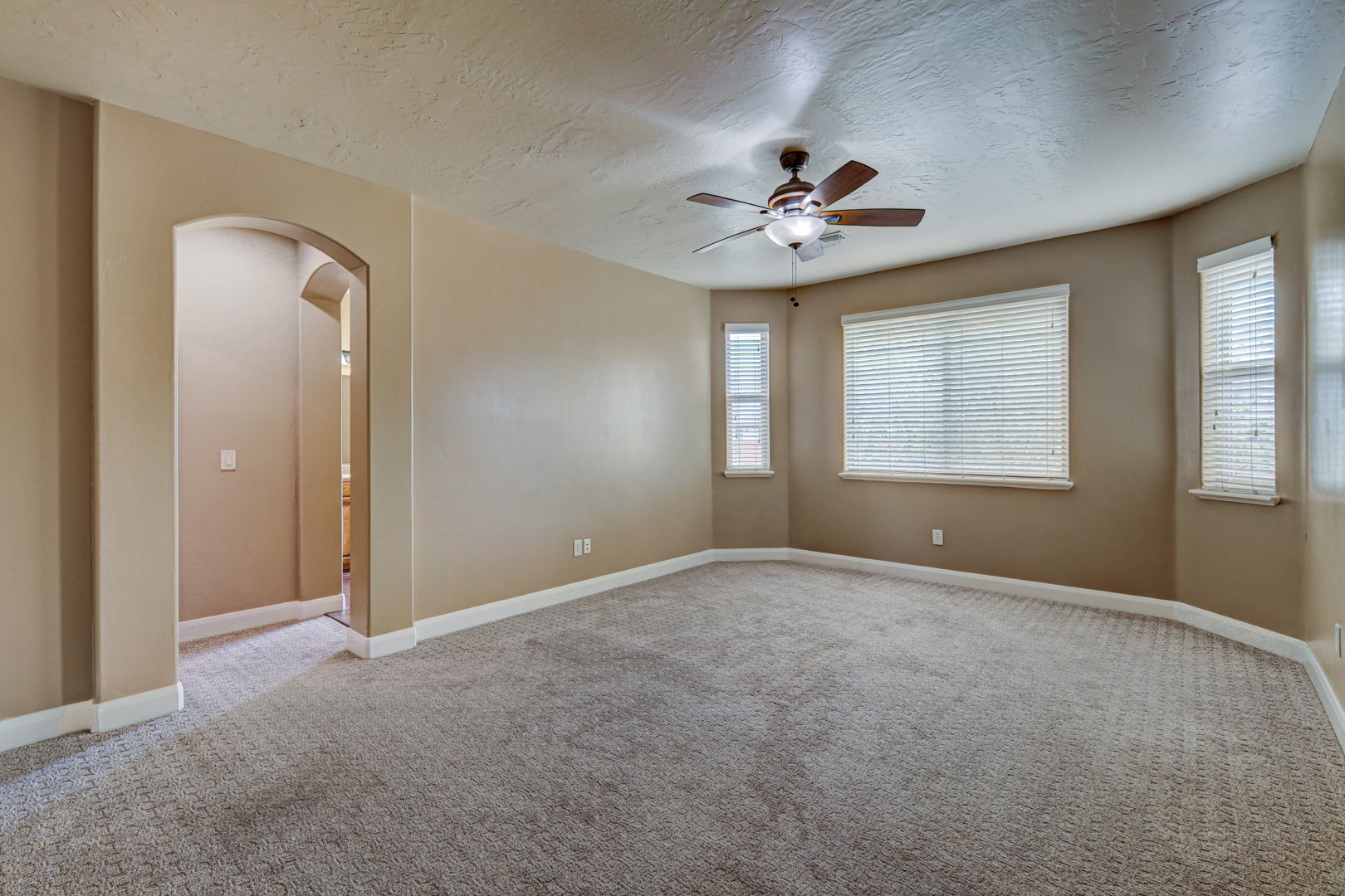 Spacious room featuring neutral carpet flooring, arched doorway, multiple windows with blinds, a ceiling fan with integrated lighting, and neutral wall paint