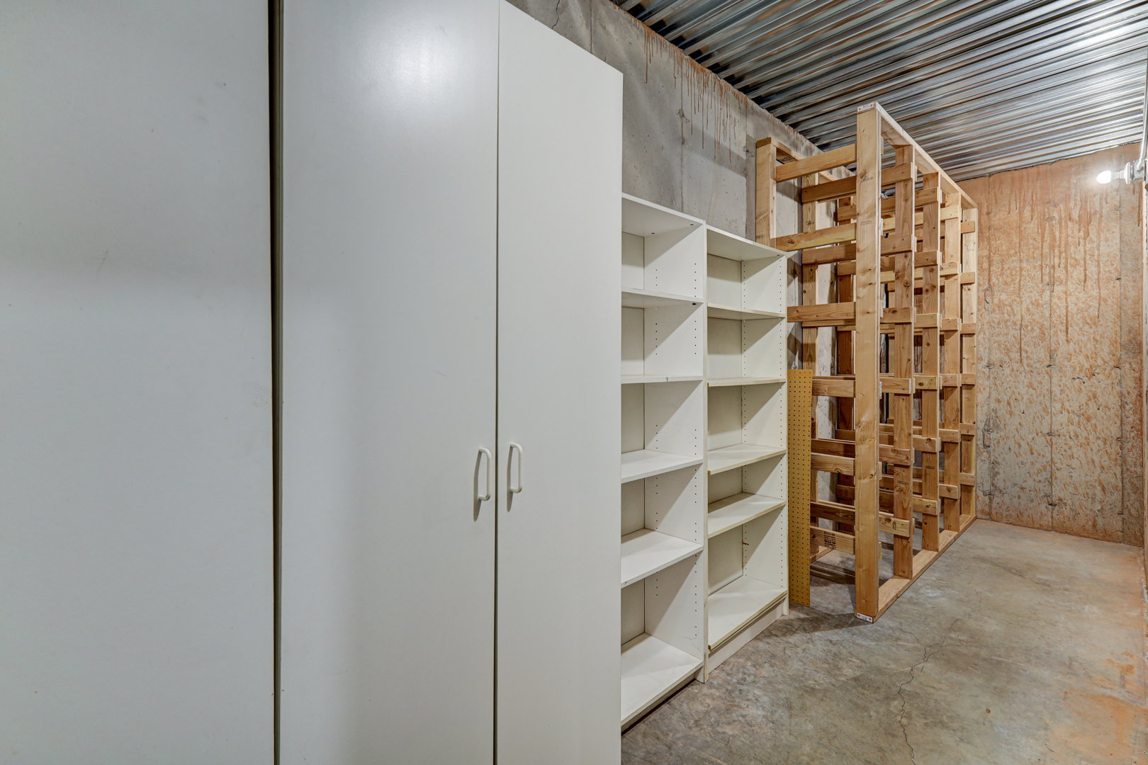 Utility room featuring concrete flooring, exposed ceiling joists, and built-in storage solutions including cabinetry, open shelving, and a wood-frame rack system