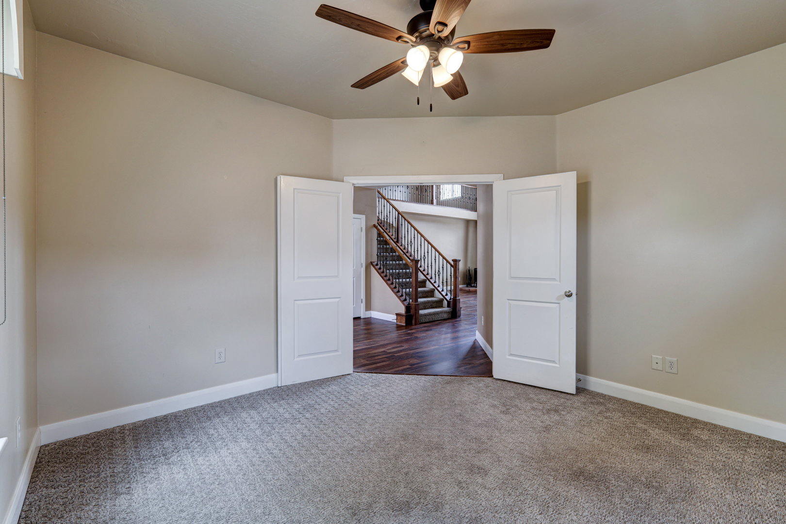Carpeted room featuring a ceiling fan with integrated lighting, painted walls, and an open doorway revealing a staircase with wood-finish flooring and balusters