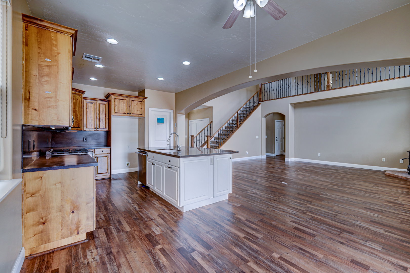 Open-concept living space featuring wood-finish flooring, a kitchen island with integrated sink, recessed lighting, a ceiling fan, and a staircase with decorative wrought iron railing