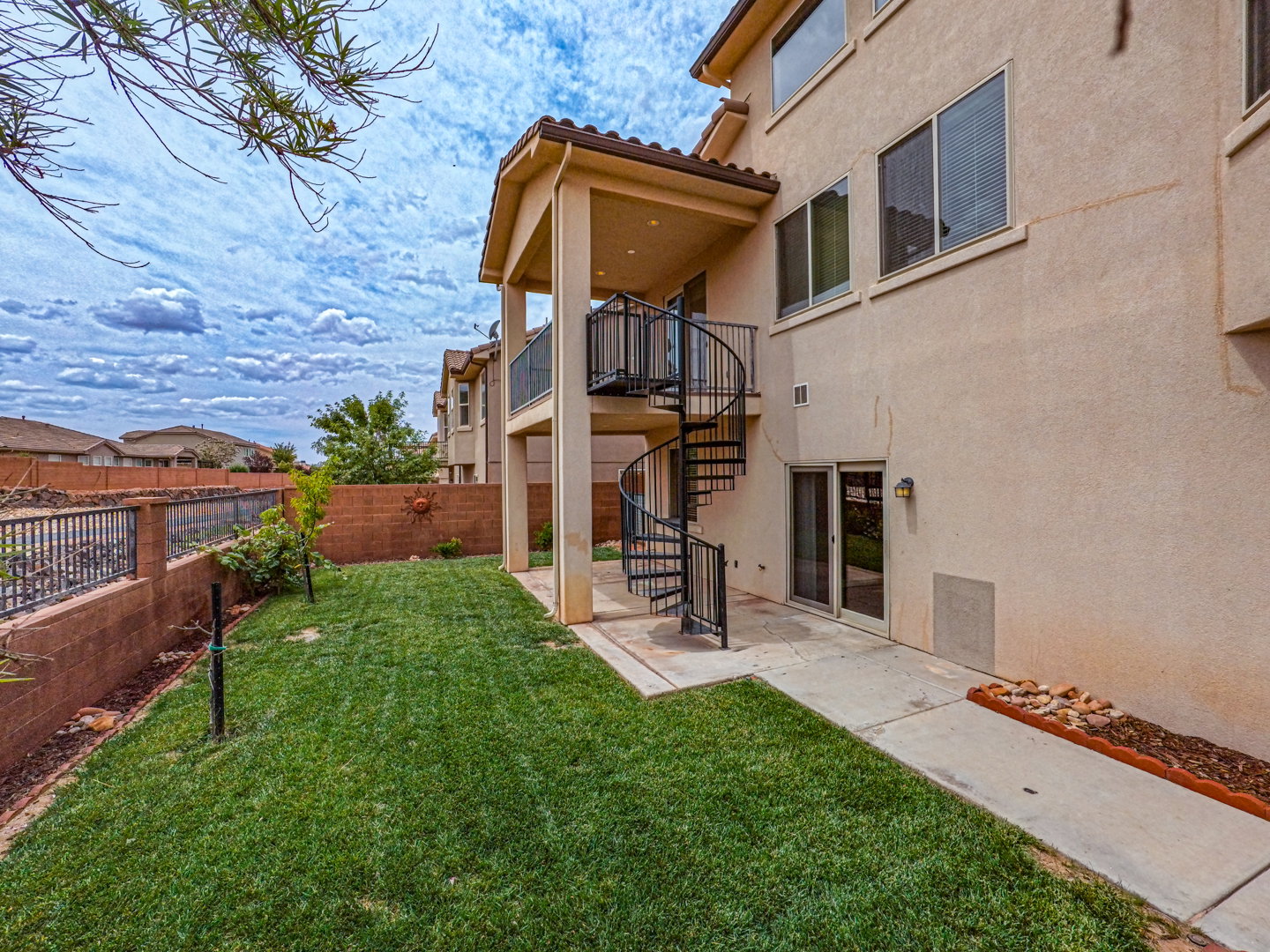 Two-story stucco exterior with a spiral staircase, covered patio, ground-level sliding glass doors, and a private yard featuring a retaining wall and wrought iron fencing