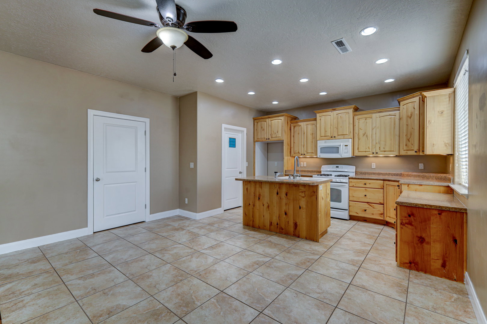 Open-concept kitchen featuring light wood cabinetry, white appliances, a central island with an integrated sink, recessed lighting, and tile flooring