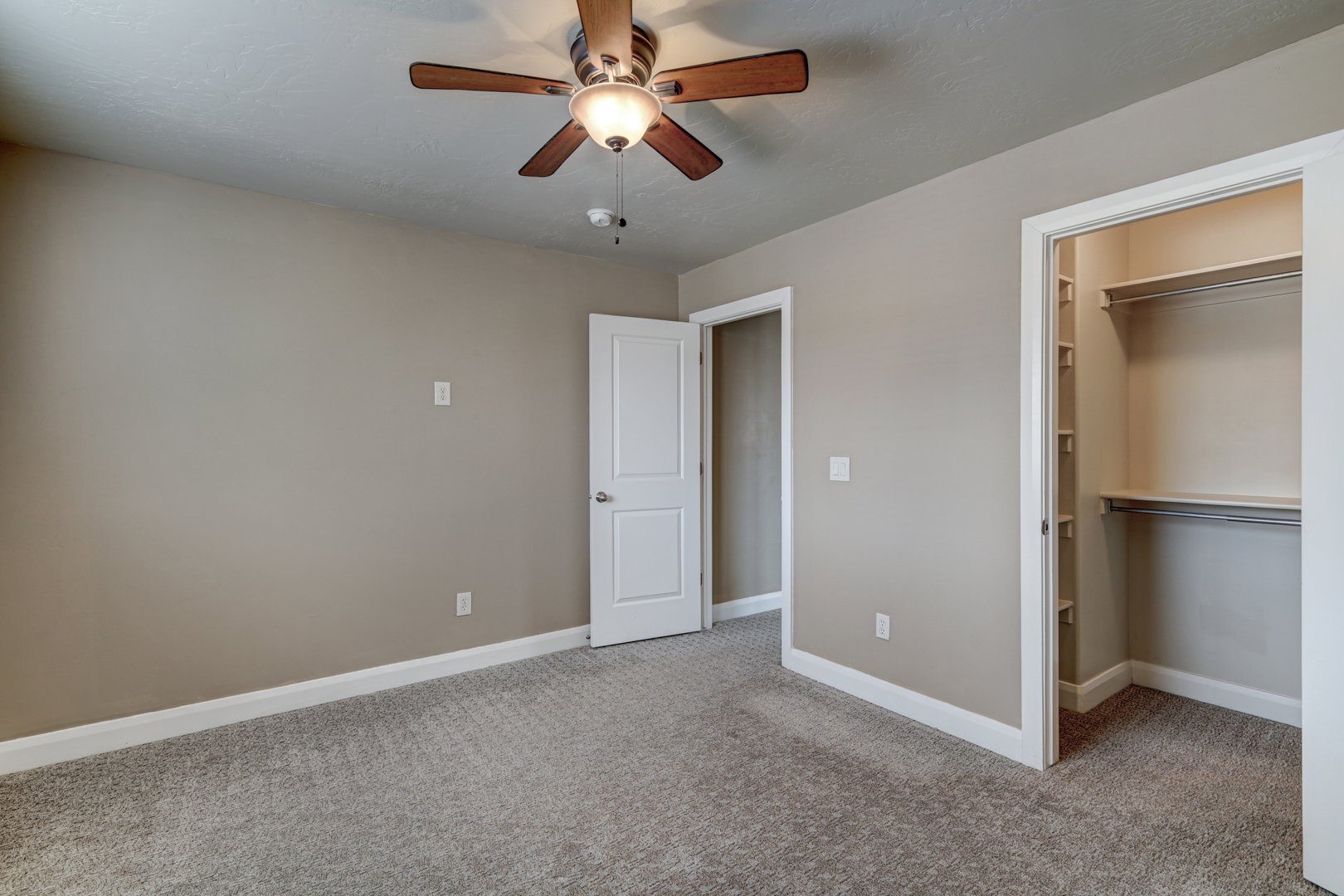 Carpeted room featuring a ceiling fan with light fixture, neutral wall paint, white trim, and a built-in closet with shelving and hanging rods