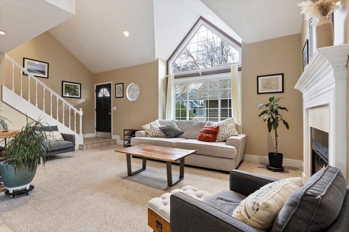 Vaulted ceilings accentuate the expansive fenestration, showcasing a fireplace with a white mantel, staircase with white balusters, and a black front entry door