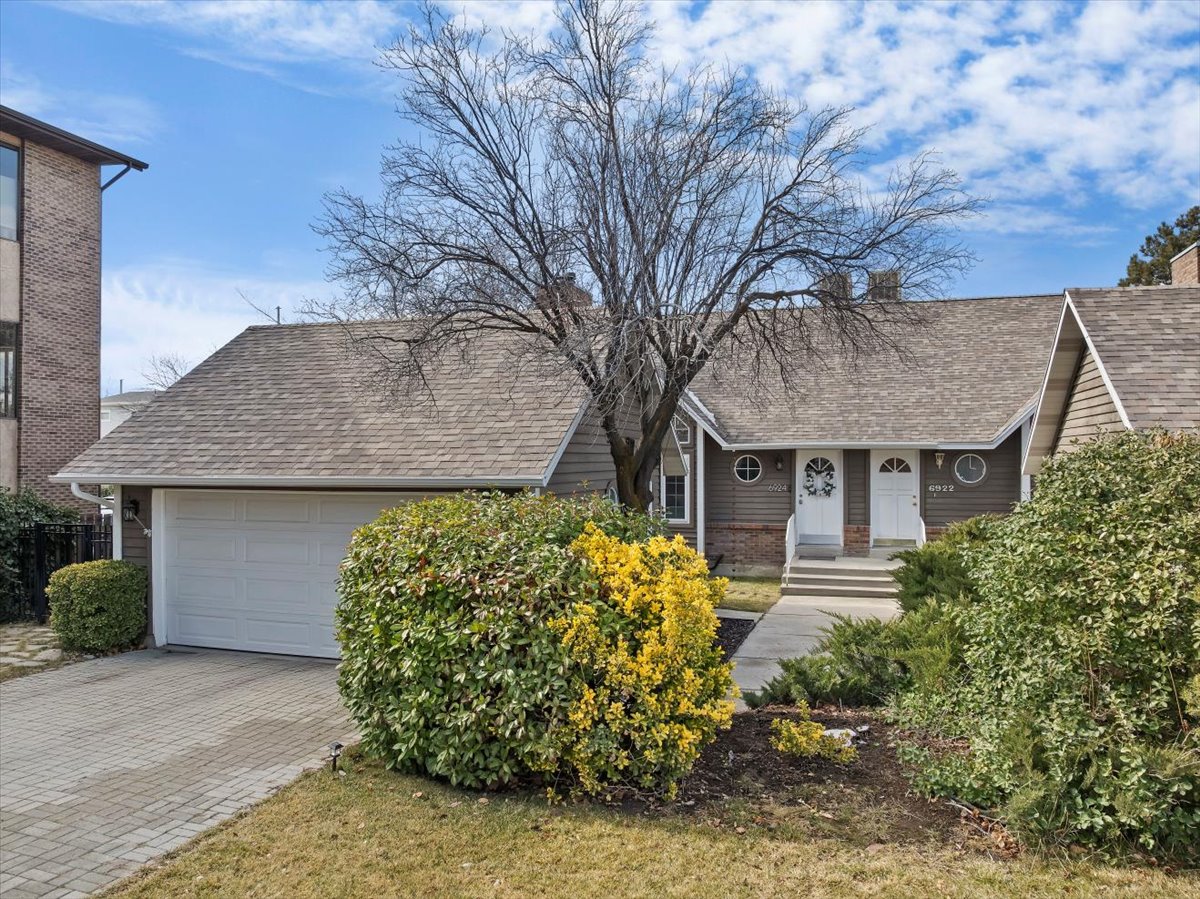 Duplex property featuring two separate entrances with white doors, a single-car garage, brick paver driveway, and varied shrubbery