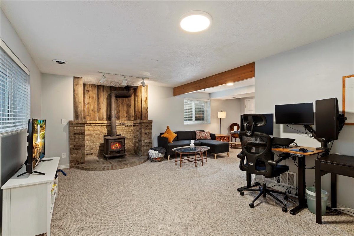 Spacious recreation room featuring a brick-base wood-burning stove with wood paneling, light-colored carpeting, a exposed wood beam, and track lighting