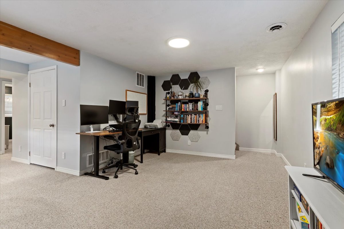 Spacious carpeted room featuring a white door, light grey walls, a ceiling-mounted light fixture, a window with blinds, and a visible wooden beam