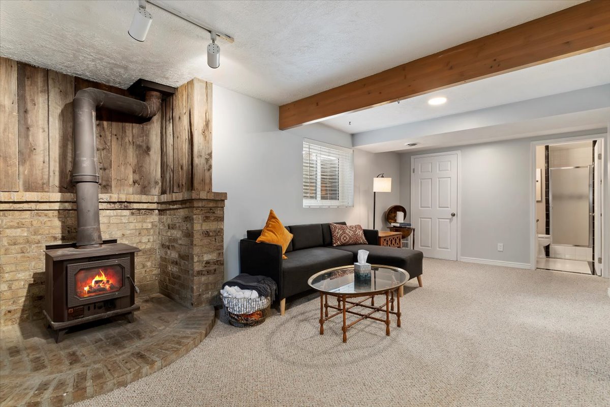 Lower level living space featuring a wood-burning stove with a brick and wood-paneled surround, track lighting, exposed wood beam, carpeted flooring, and a window with blinds
