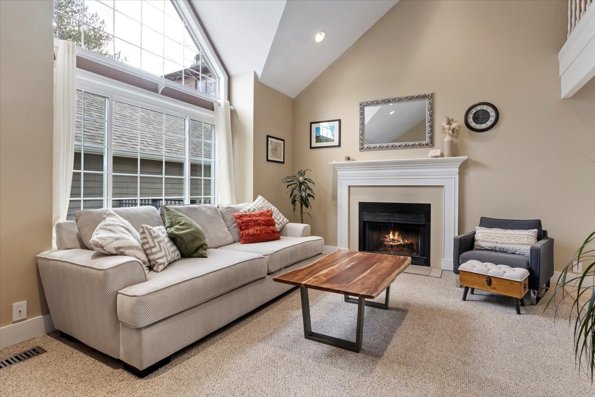 Living area featuring a floor-to-ceiling window arrangement with transom windows, vaulted ceilings, a fireplace with a white mantel, and neutral carpeting