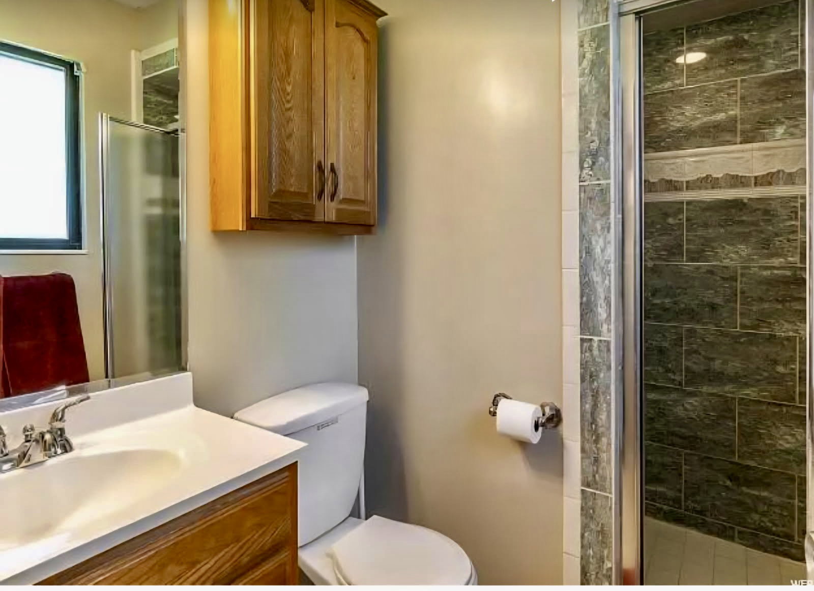 Bathroom featuring a shower with tiled surround, wood-finish vanity with integrated sink, and an overhead wood-finish cabinet
