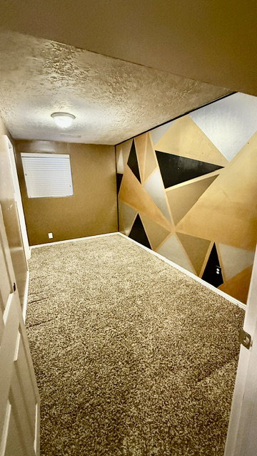 Interior room featuring textured carpet flooring, a geometric accent wall with gold, black, and silver tones, and a window with horizontal blinds