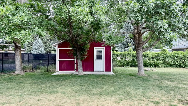 Crimson storage shed with white trim, a white door, and a concrete foundation, set within a grassy area featuring mature trees