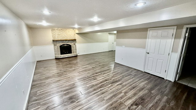 Spacious room featuring wood-finish flooring, a corner brick fireplace with a wood mantel, white wainscoting, and recessed lighting