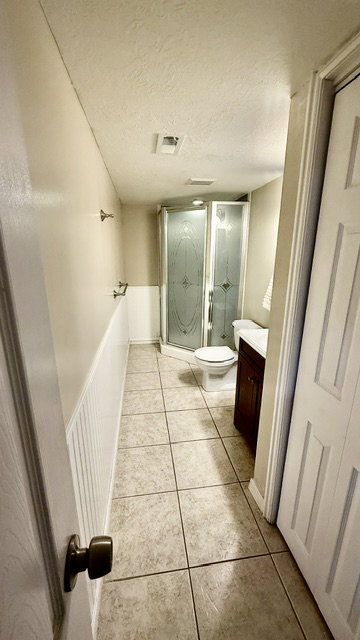 Corner shower enclosure with decorative glass panels, tile flooring, a dark wood-finish vanity, and wainscoting