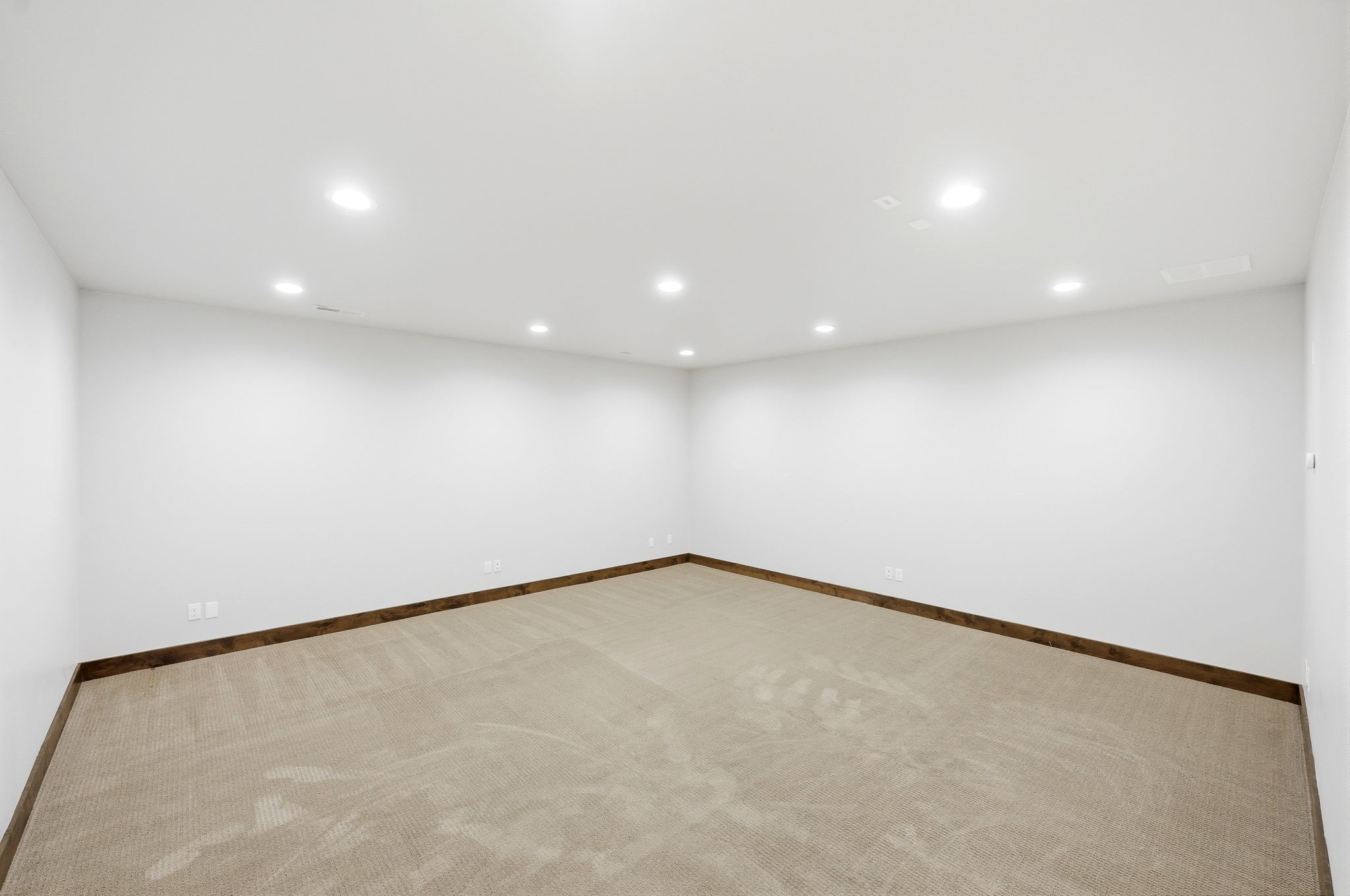 Spacious room with white walls, recessed ceiling lighting, light-toned carpet flooring, and dark wood-finish baseboards