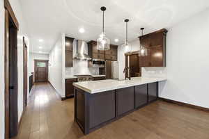 Open-concept kitchen featuring wood-finish flooring, dark wood cabinetry, a large kitchen island with a light-toned countertop, stainless steel appliances, and pendant lighting