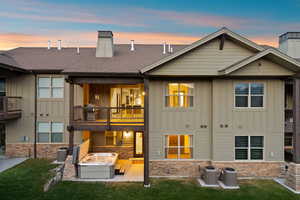 Rear exterior featuring a covered deck, lower patio with hot tub, and stone accent base