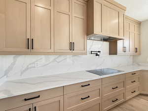 Kitchen featuring extensive light wood-finish cabinetry with black hardware, white countertops and backsplash with grey veining, a built-in cooktop, and an integrated range hood