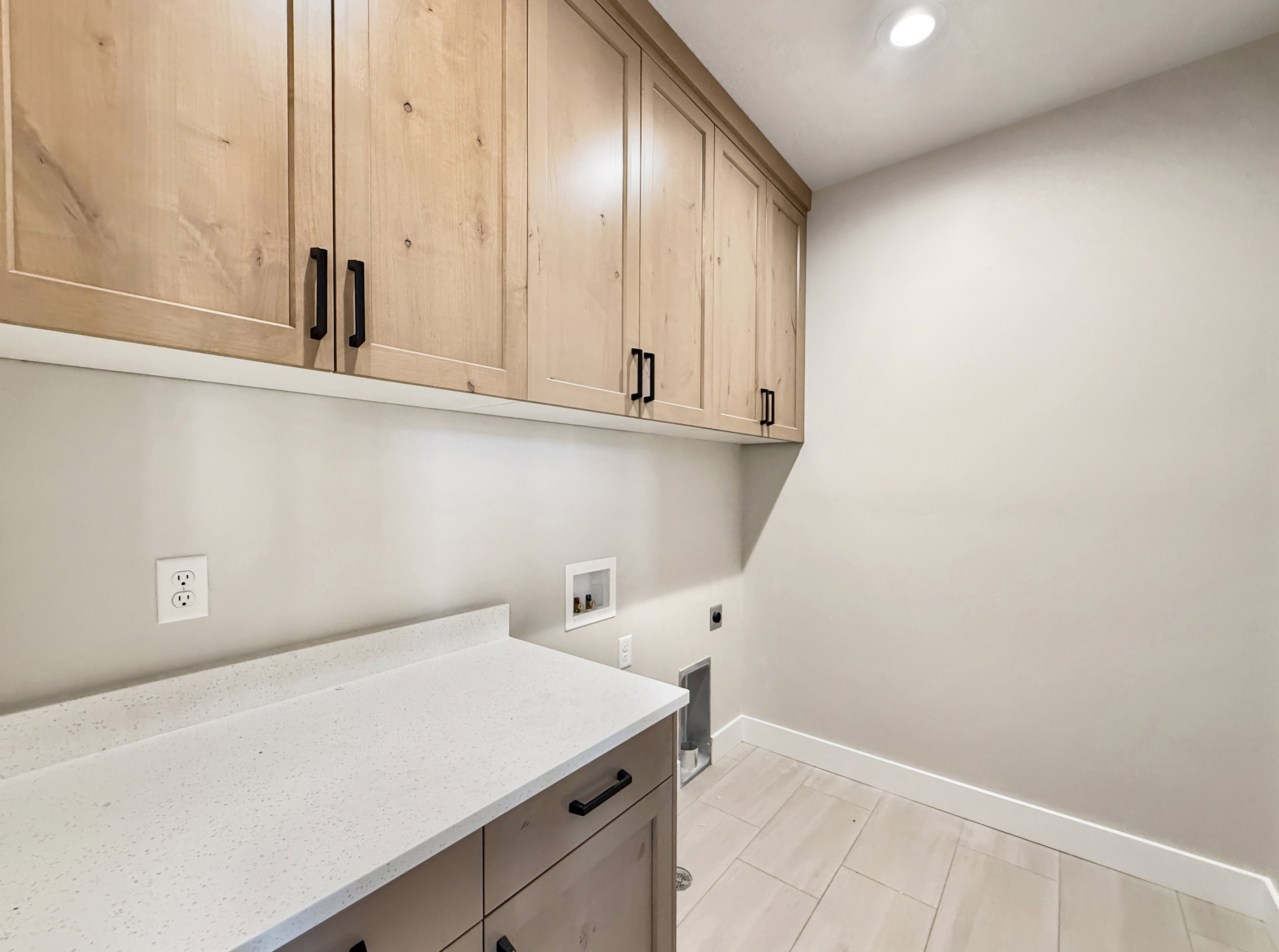 Laundry area featuring light wood-finish cabinetry with matte black hardware, a light speckled countertop, and light-toned tile flooring