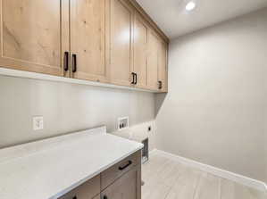 Laundry area featuring light wood-finish cabinetry with matte black hardware, a light speckled countertop, and light-toned tile flooring