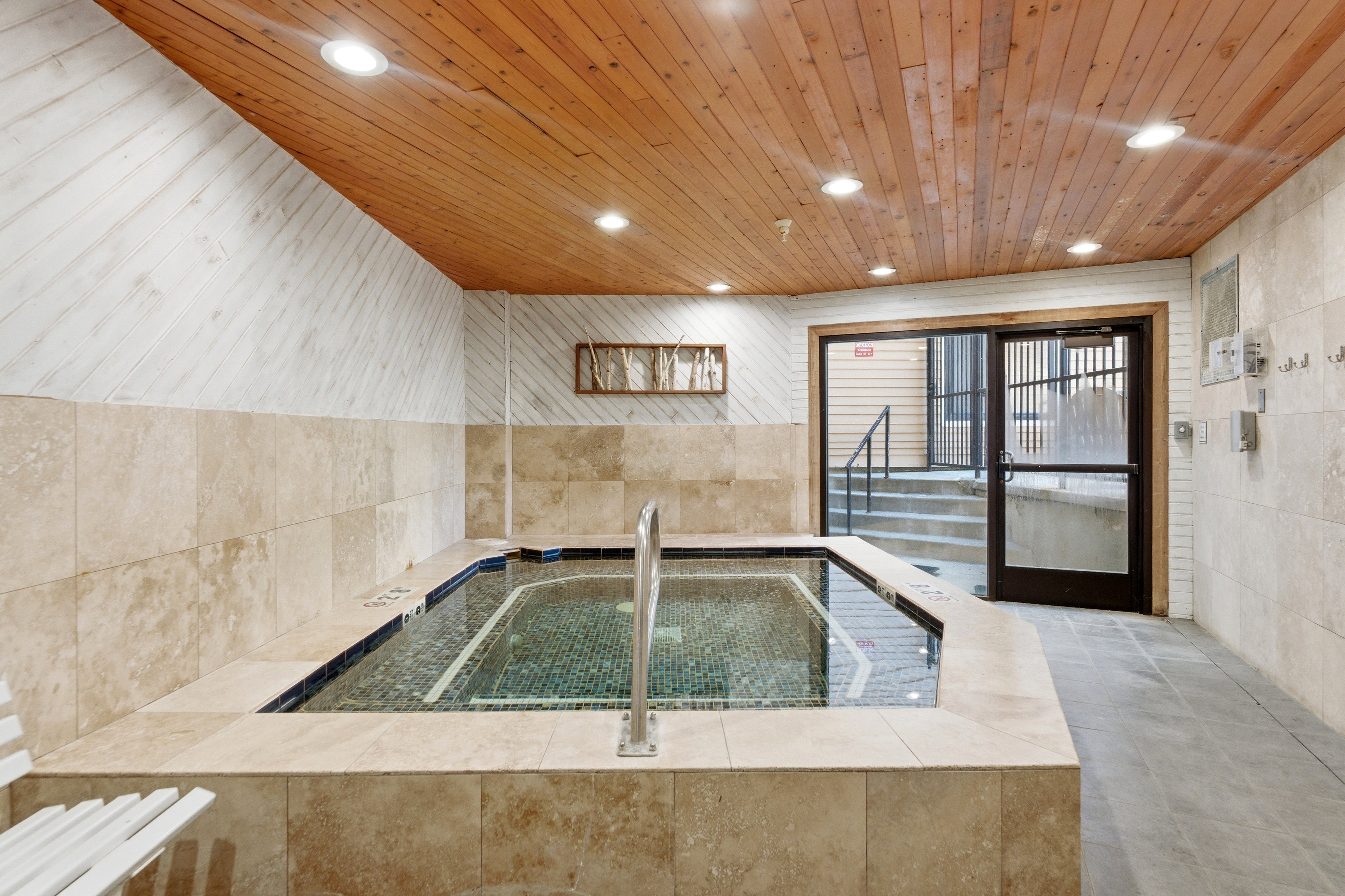 Indoor hot tub with mosaic tile interior, surrounded by light-toned tile decking and walls