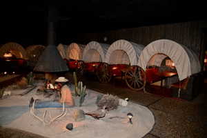 Western-themed indoor dining area featuring multiple covered wagons with built-in tables and seating, a central freestanding fireplace, desert landscaping with artificial cacti, and a textured wall paneling
