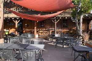 Outdoor dining area featuring multiple metal tables and chairs, overhead red sun shades with string lighting, and a rustic wood-sided building with exposed beam details