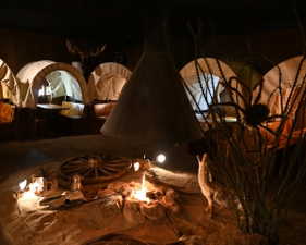 Indoor immersive space featuring multiple covered wagon structures, a central simulated campfire with surrounding rocks, a wagon wheel element, and a large decorative hood
