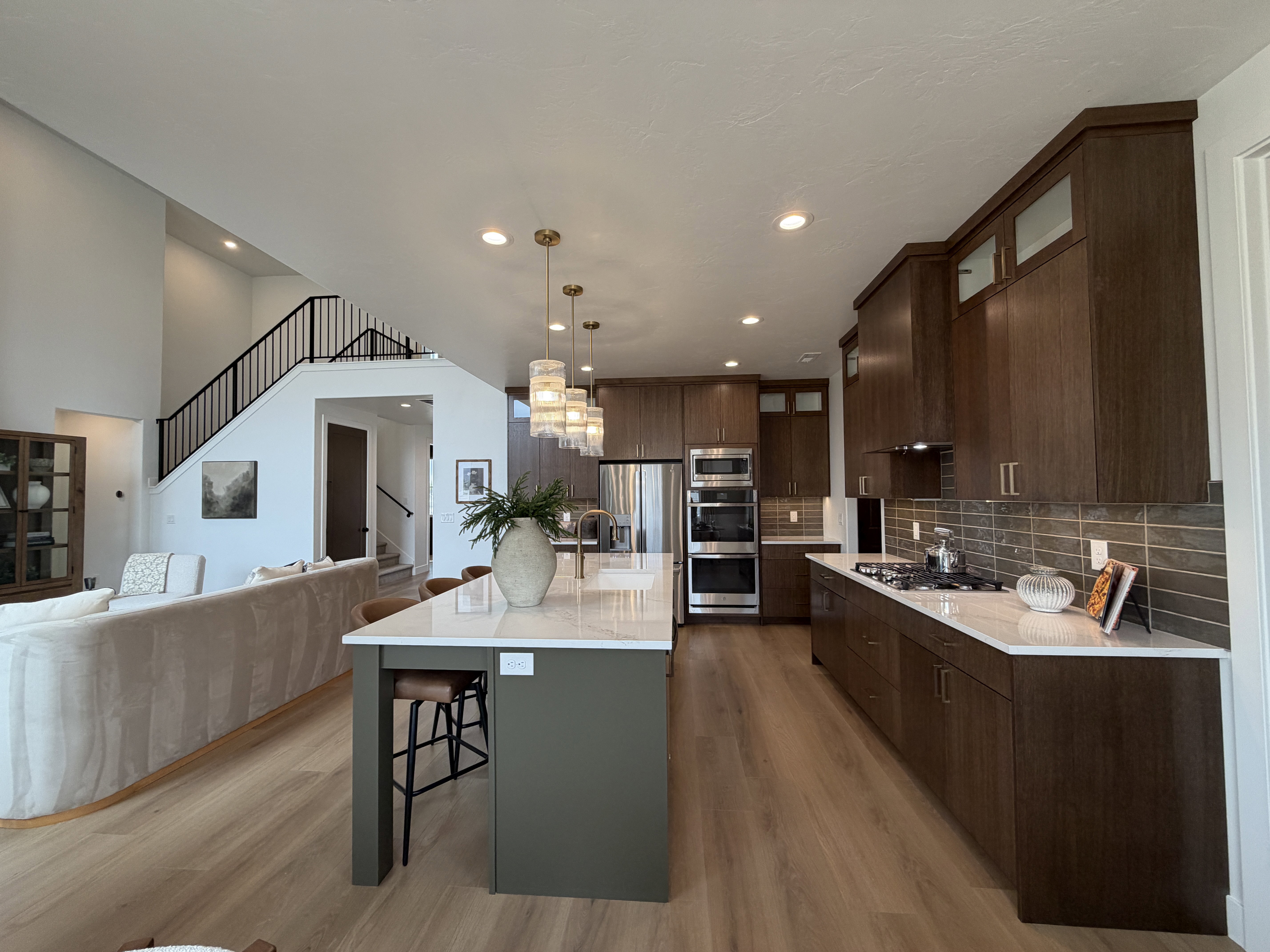 Open-concept kitchen and living area featuring wood-finish flooring, a kitchen island with seating, dark wood-tone cabinetry, stainless steel appliances, and a staircase with black metal railings
