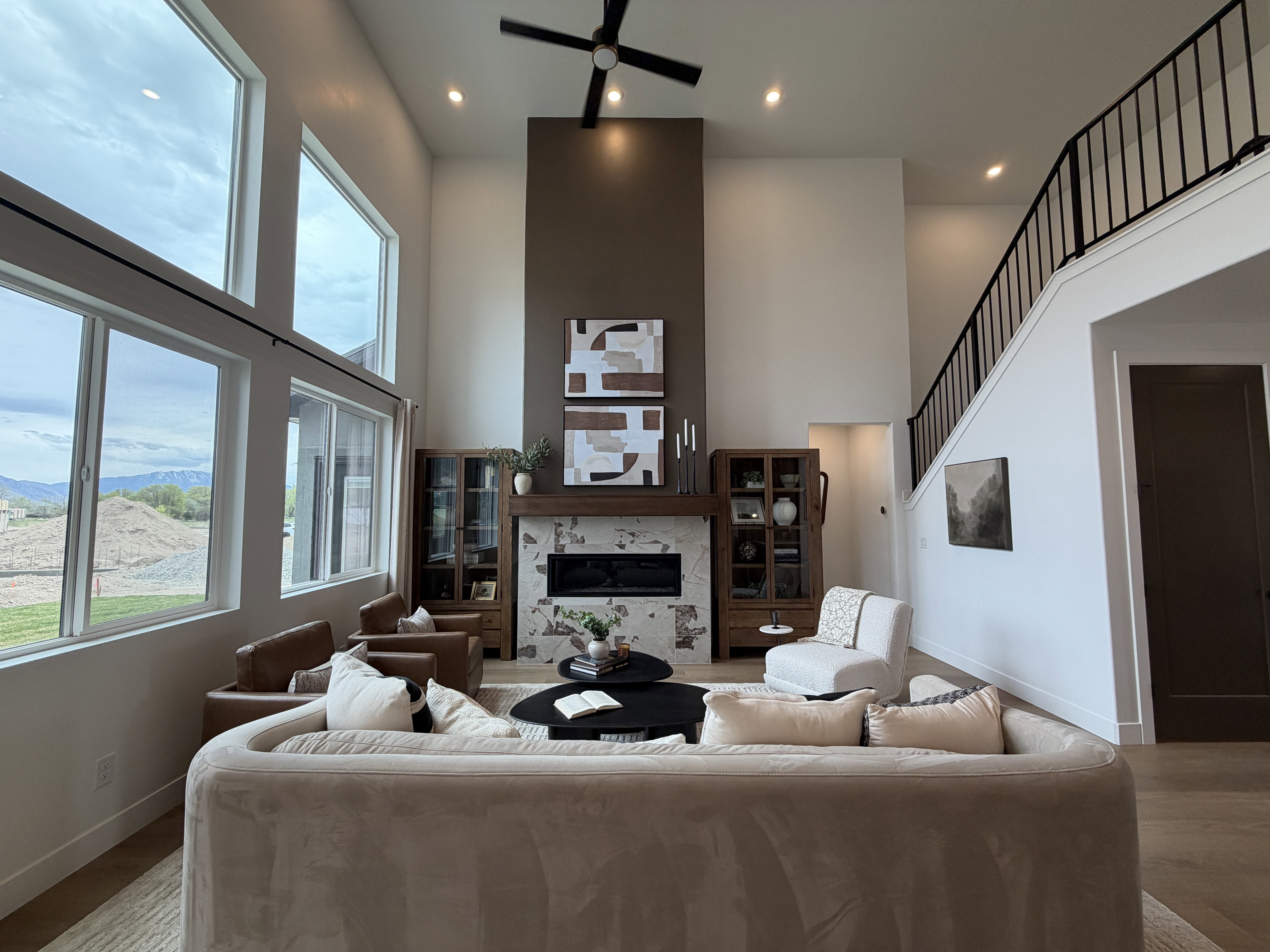 Vaulted great room with floor-to-ceiling windows, modern fireplace with stone surround and wood mantle, contemporary ceiling fan, recessed lighting, and a black metal staircase railing