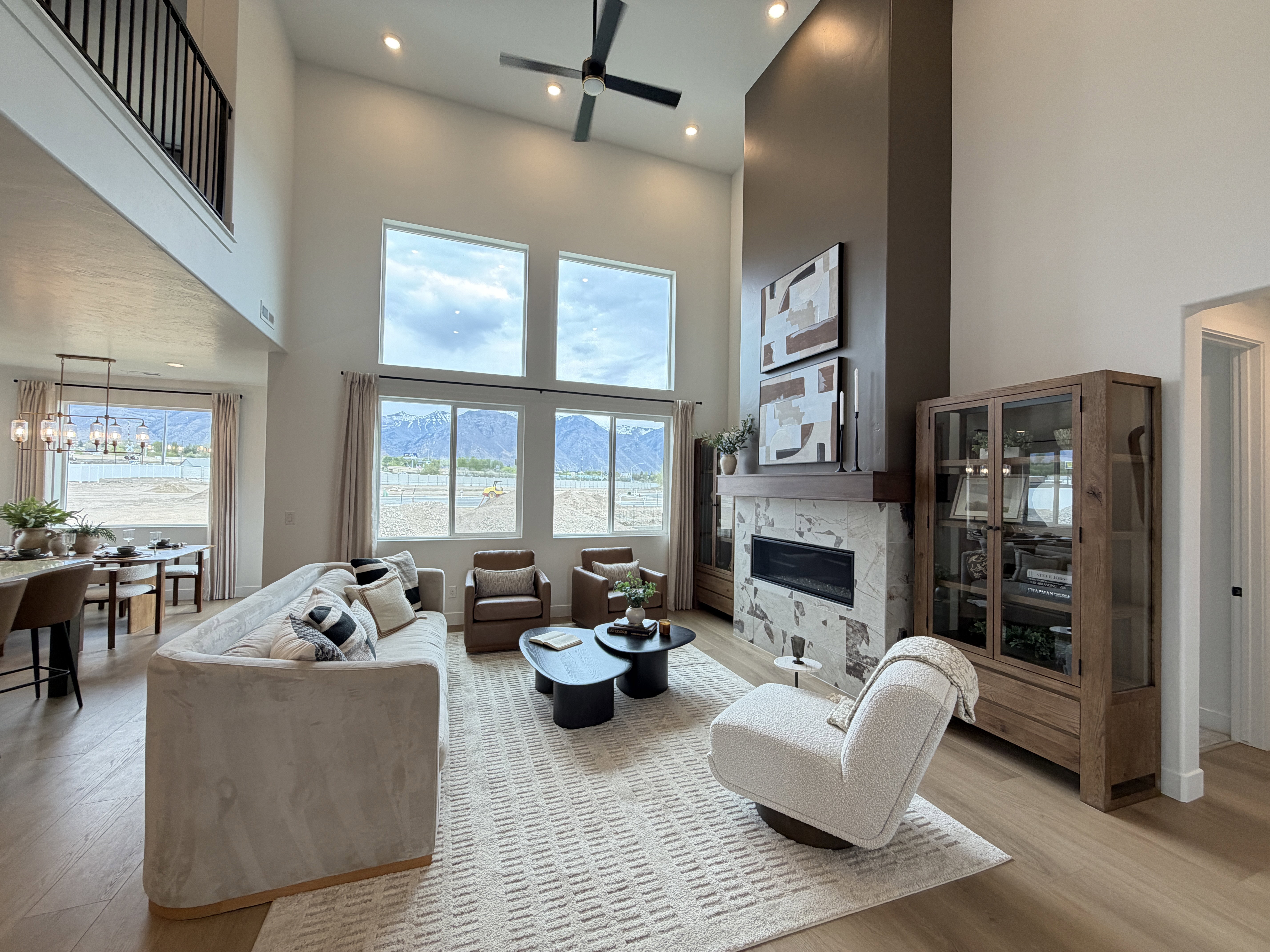 Two-story living room featuring expansive floor-to-ceiling windows, a stone-clad fireplace with a dark accent column, a second-story interior balcony, and wood-finish flooring