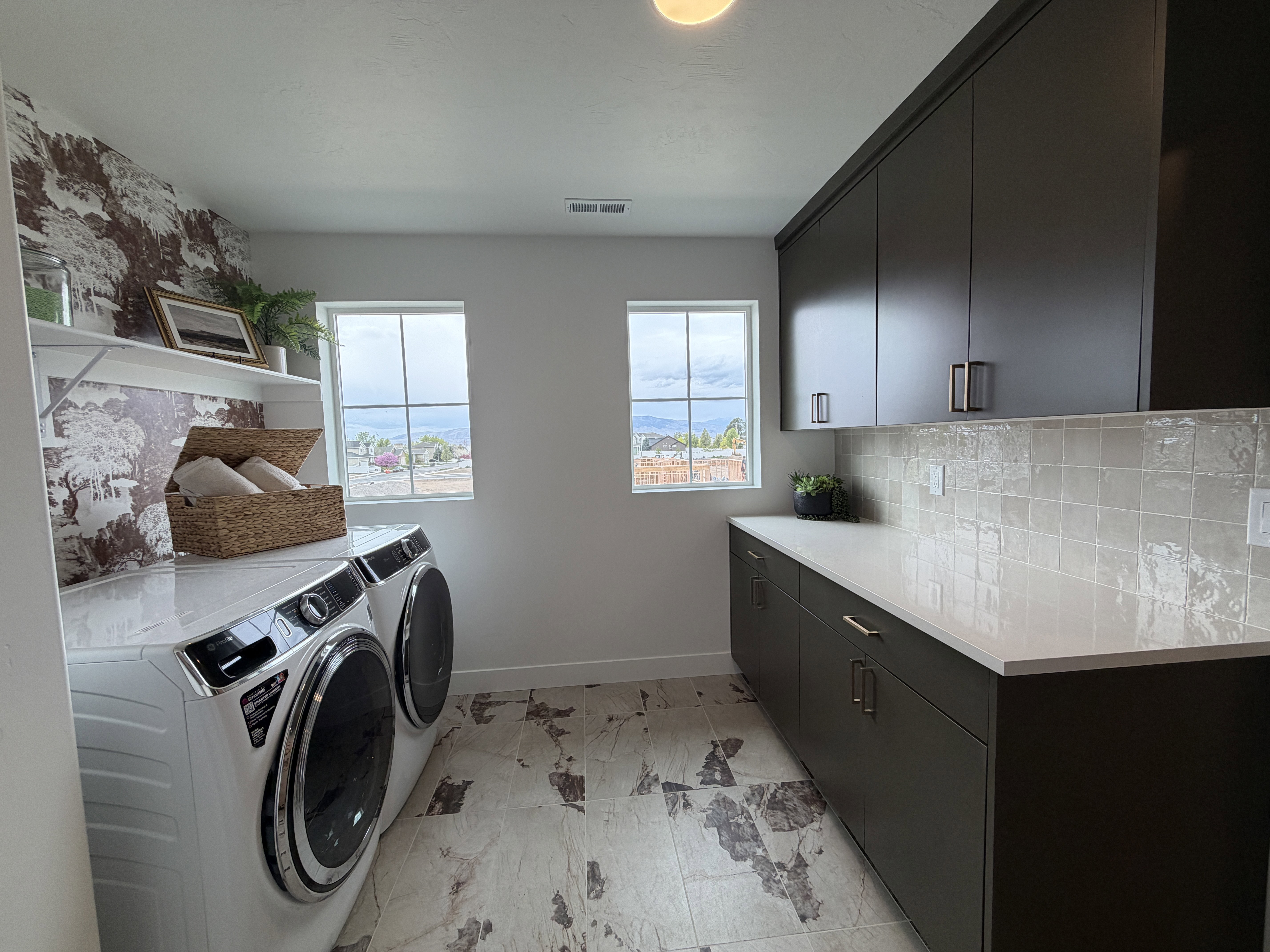 Dedicated laundry area featuring a patterned accent wall with open shelving, a built-in countertop, and dark cabinetry with modern hardware