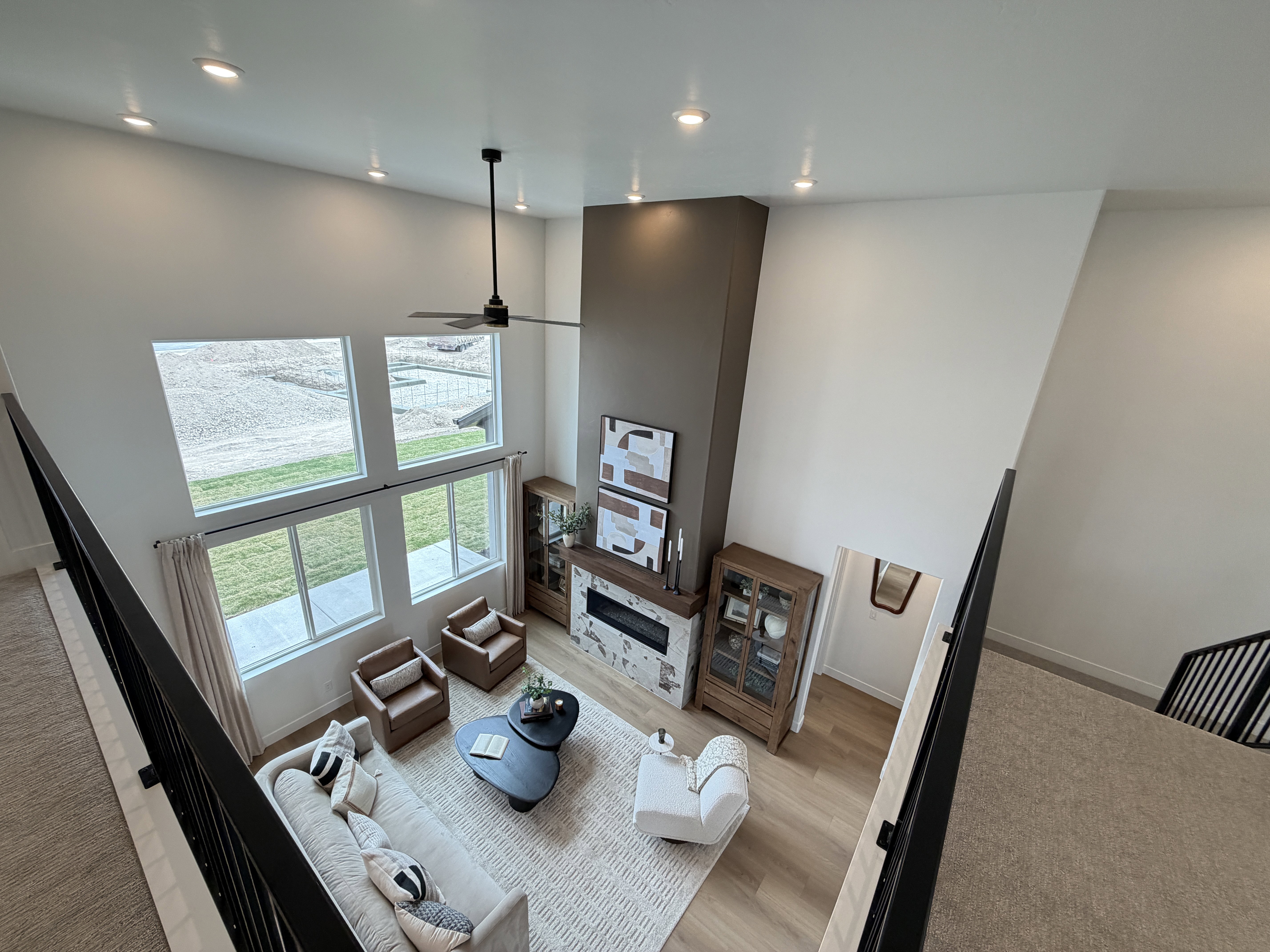 Spacious living area featuring a two-story ceiling, a multi-pane window configuration, wood-finish flooring, a modern fireplace with a white stone surround, and a ceiling fan with integrated lighting