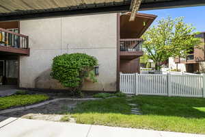 Private patio area with stucco exterior and white picket fencing, featuring a mature shrub, integrated wall lighting, and landscaping